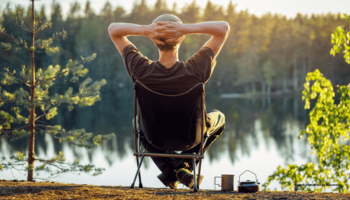 Das Foto zeigt einen Mann auf einem Campingstuhl sitzend vor einem See mit strahlender Sonne.