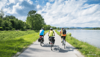 Das Foto zeigt drei Personen auf einem Radweg bei schönem Wetter fahrend in der Natur bei einem Fluss.