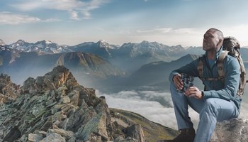 Das Foto zeigt einen Mann, welcher auf einem Berg sitzt und ein Fernglas hält.