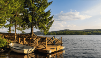 Es ist ein kleines, weißes Boot neben einem Holzsteg im Wasser zu sehen. Der Steg führt an Land, wo mehrere grüne Bäume stehen. Im Vordergrund und Hintergrund des Bildes ist Wasser zu sehen. In der Distanz sieht man eine hügelige Waldlandschaft. Der Himmel ist blau.