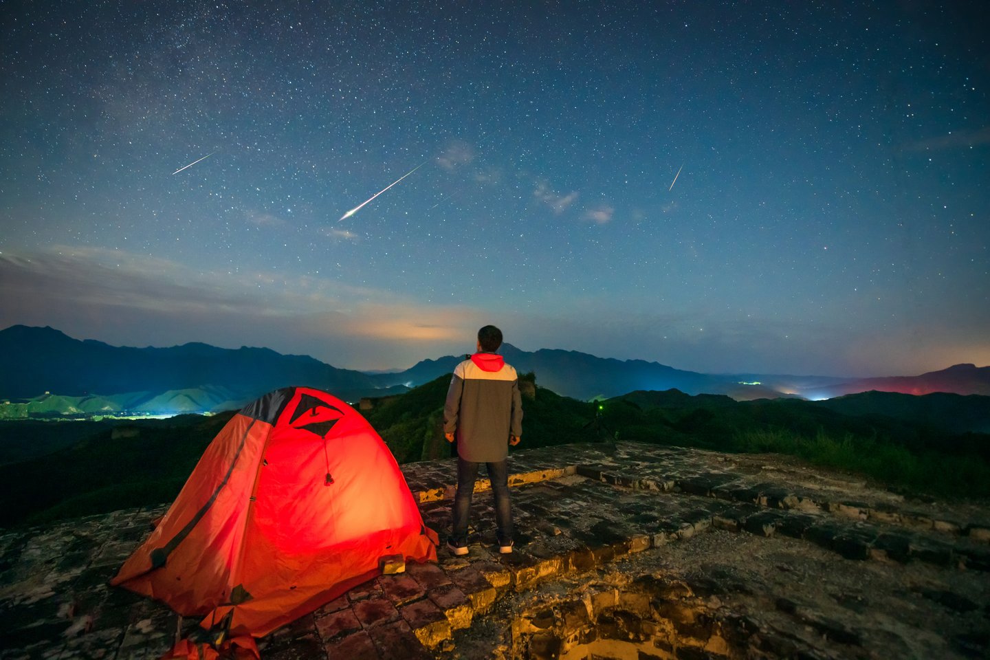 Das Foto zeigt einen Mann, der vor seinem Zelt steht und im nächtlichen Sternenhimmel Sternschnuppen beobachtet. 