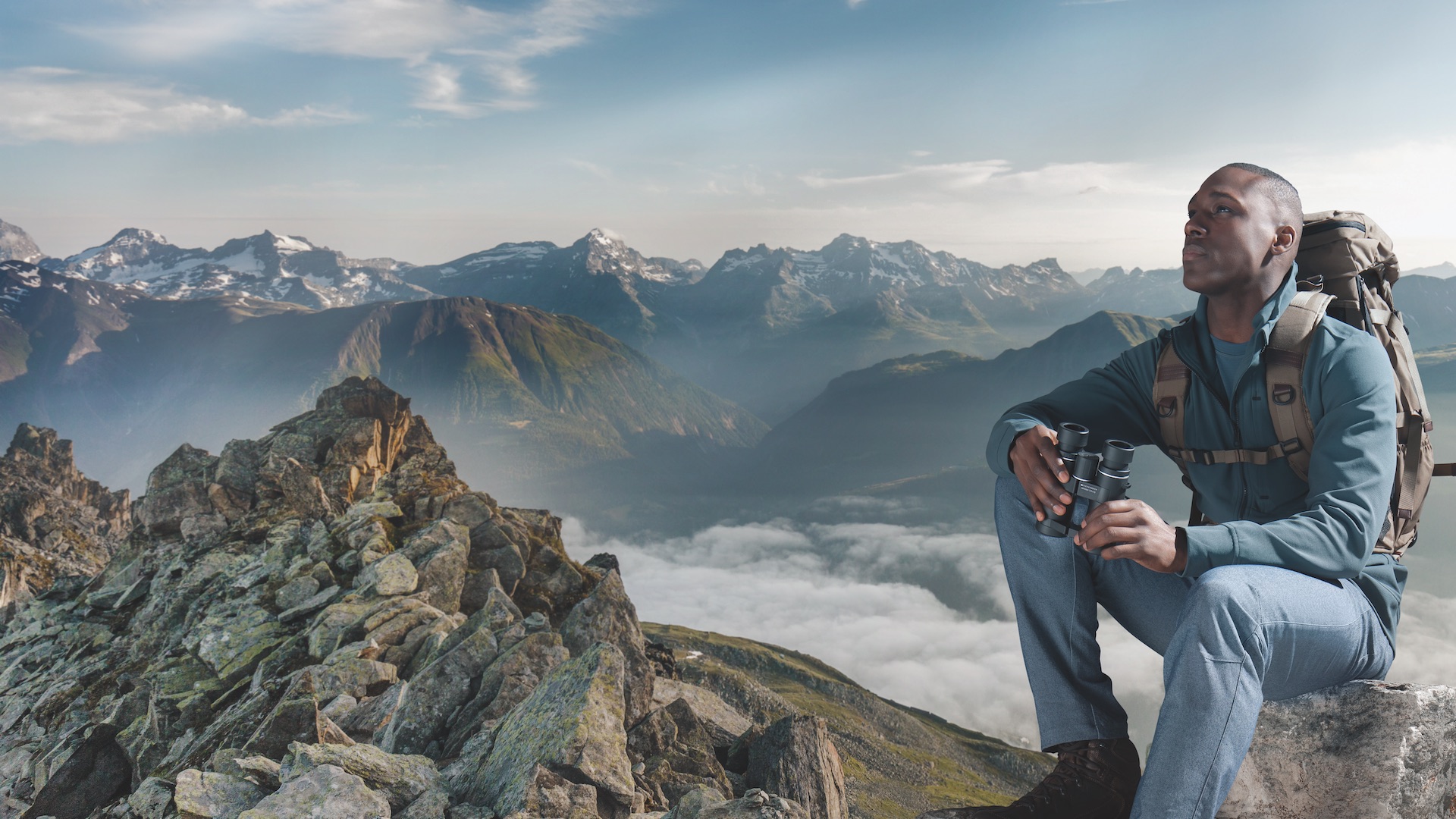 Das Foto zeigt einen Mann, welcher auf einem Berg sitzt und ein Fernglas hält.